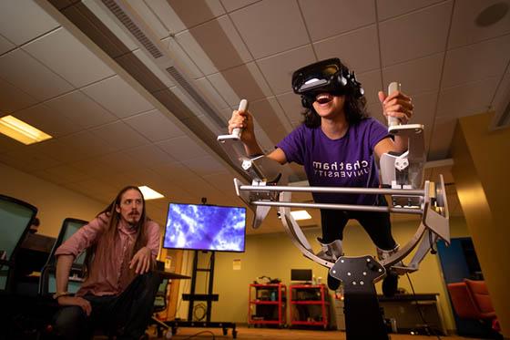 Photo of a student wearing a purple Chatham University shirt, 在虚拟现实机里, 而一位教授在旁边看着